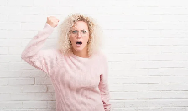 Young blonde woman with curly hair over white brick wall angry and mad raising fist frustrated and furious while shouting with anger. Rage and aggressive concept.