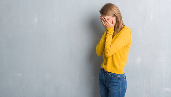 Beautiful young woman standing over grunge grey wall with sad expression covering face with hands while crying. Depression concept.