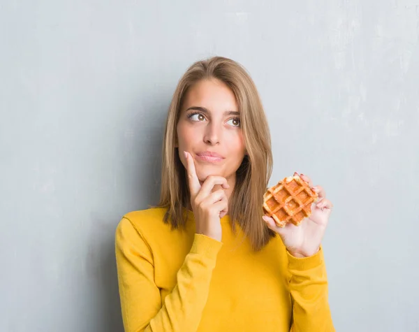 美丽的年轻女人在垃圾灰墙上吃华夫饼严肃的脸思考问题 非常困惑的想法 — 图库照片