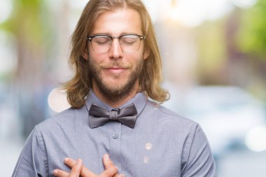 Göğüs kapalı gözler ve yüzünde minnettar jest ile gülümseyen izole arka plan üzerinde papyon giyiyor uzun saçlı yakışıklı delikanlı eller. Sağlık kavramı.