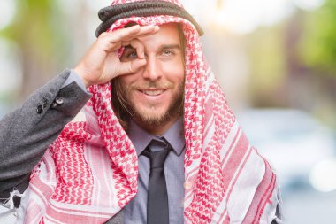 Genç yakışıklı Arap adam uzun saçlı gülümseyen, parmakları ile mutlu yüz üzerinden seyir göz el ile ok jest yapıyor izole arka plan üzerinde keffiyeh giyiyor.