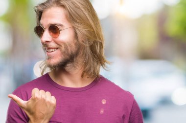 Seyir ve başparmak yanında yukarıyı mutlu yüz ile gülümseyen izole arka plan üzerinde güneş gözlüğü takmış uzun saçlı yakışıklı delikanlı.