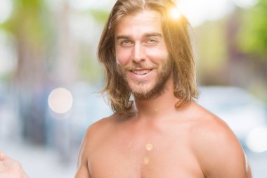 Genç yakışıklı gömleksiz adam uzun saçlı seksi vücut hayrete ve el ile sunulması ve parmak ile işaret ederken kameraya gülümseyen izole arka plan üzerinde gösterilen.