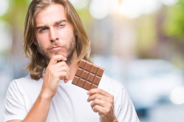 Uzun saç beslenme ile yakışıklı delikanlı çikolata bar izole arka plan ciddi bir yüz düşünme hakkında soru, çok karışık bir fikir üzerinde