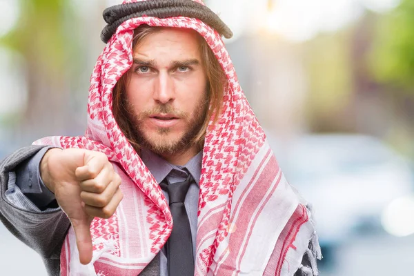 Genç yakışıklı Arap adam uzun saçlı mutsuz ve kızgın ret gösterilen ve başparmak aşağı hareketi ile negatif seyir izole arka plan üzerinde keffiyeh giyiyor. Kötü ifade.