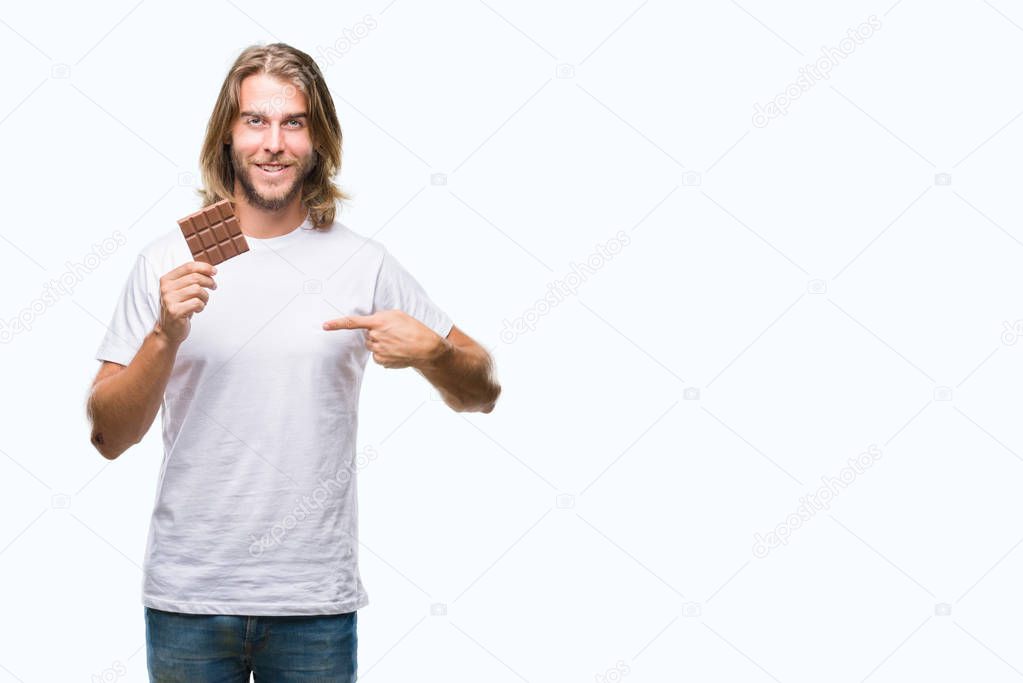 Young handsome man with long hair eating chocolate bar over isolated background with surprise face pointing finger to himself