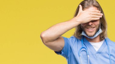 Genç yakışıklı Doktor adam gülümsüyor ve gözleri için sürpriz kapsayan yüz el ile gülüyor izole arka plan üzerinde uzun saçlı. Kör kavramı.