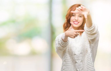 İzole arka plan elle yapma çerçeve ve parmakları ile mutlu yüz gülümseyen kış kazak giyen genç güzel kadın. Yaratıcılık ve Fotoğrafçılık kavramı.