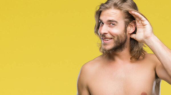 Young handsome shirtless man with long hair showing sexy body over isolated background smiling with hand over ear listening an hearing to rumor or gossip. Deafness concept.