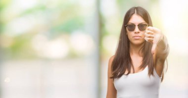 Genç güzel İspanyol mutsuz ve kızgın ret gösterilen ve başparmak aşağı hareketi ile negatif seyir güneş gözlüğü takıyor. Kötü ifade.