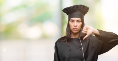 Young hispanic cook woman wearing chef uniform with angry face, negative sign showing dislike with thumbs down, rejection concept clipart