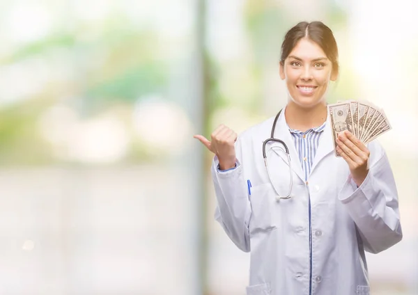 Joven Doctora Hispana Sosteniendo Dólares Señalando Mostrando Con Pulgar Hacia — Foto de Stock