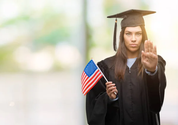 Young hispanic woman wearing graduated uniform holding flag of america with open hand doing stop sign with serious and confident expression, defense gesture