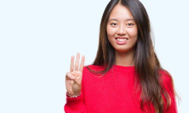 Genç Asyalı kadın giyiyor kış kazak gösterilen ve parmakları ile işaret izole arka plan üzerinde üçüncü ise kendinden emin ve mutlu gülümseyerek.