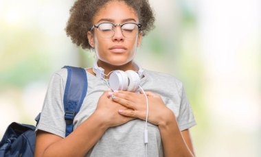 Göğüs kapalı gözlerle ellerinde ve yüzünde minnettar jest ile gülümseyen izole arka plan üzerinde kulaklık ve sırt çantası giyen genç afro Amerikan öğrenci kadın. Sağlık kavramı.