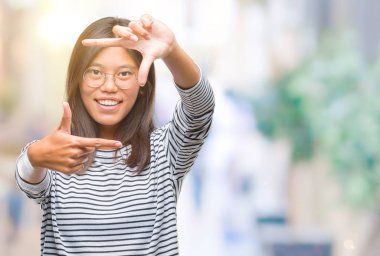 Genç Asyalı kadın elle yapma çerçeve ve parmakları ile mutlu yüz gülümseyen izole arka plan üzerinde gözlük takıyorum. Yaratıcılık ve Fotoğrafçılık kavramı.