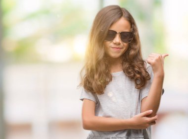 Esmer Latin kız arıyorum ve başparmak yanında yukarıyı mutlu yüz ile gülümseyen güneş gözlüğü takıyor.