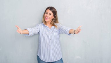 Gri grunge duvar sarılmak için kollarını açarak gülümseyerek kameraya bakarak moda iş kıyafeti giymiş genç yetişkin kadın. Neşeli ifade kucaklayan mutluluk.