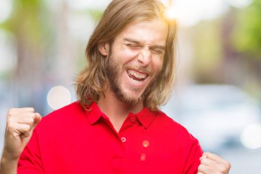 Gülümseyen ve başarı için çığlık yakışıklı delikanlı izole arka plan çok mutlu ve heyecanlı kaldırdı, silah ile kazanan jest yapıyor üzerinde uzun saçlı. Kutlama kavramı.