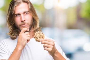 Young handsome man with long hair eating chocolate cooky over isolated background serious face thinking about question, very confused idea clipart