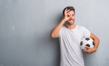 Mutlu yüz el parmaklarının arasından seyir göz üzerinde ok işaretiyle yapıyor gülümseyen ile futbol futbol topu tutan gri grunge duvarın üzerinden yakışıklı genç