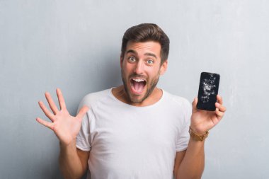 Yakışıklı genç gri grunge üzerinde duvar kırık smartphone ekran çok mutlu ve heyecanlı, kocaman bir gülümseme ile çığlık ve ellerini kaldırdı kazanan ifade kutluyor zafer gösterilen