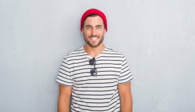 Yakışıklı genç hipster adam yüzünde lacivert t-shirt ve mutlu ve güzel bir gülümseme ile Yün şapka giyen gri grunge duvarın üzerinden. Şanslı kişi.