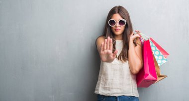 Grunge gri duvar açık eliyle dur işareti ile ciddi ve kendine güvenen ifade, savunma jest yapıyor renkli alışveriş torbaları tutan genç esmer kadın