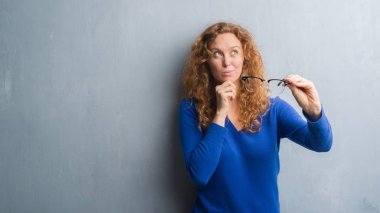 Gri grunge duvar optik gözlük ciddi bir yüz düşünme hakkında soru, çok karışık fikir tutan genç Kızıl saçlı kadın