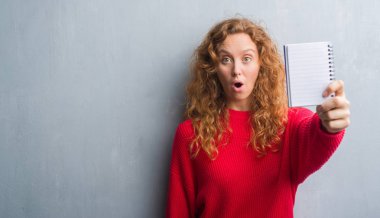 Gri grunge duvar gösteren boş defter bir sürpriz yüz, korkuyor ve heyecanlı korku ifadesi ile şok korkmuş genç Kızıl saçlı kadın