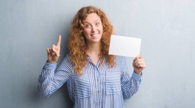 Gri grunge genç Kızıl saçlı kadın boş kartı sürpriz bir fikir ile tutarak duvar veya gülen yüz, bir numara ile işaret parmağı soru