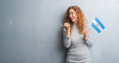 Arjantin bayrağı tutarak gri grunge duvarın üzerinden işaret ve başparmak kadar mutlu yüz gülümseyen tarafı ile gösterilen genç Kızıl saçlı kadın