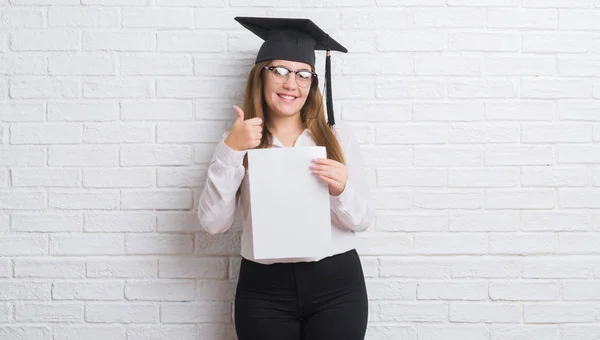 穿白砖墙的年轻成年妇女戴着研究生帽持有学位快乐的大微笑做 拇指与手指 优秀的标志 — 图库照片