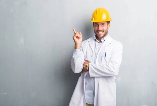 英俊的年轻工程师人在灰色的垃圾墙上戴安全帽非常高兴地指着手和手指的侧面 — 图库照片