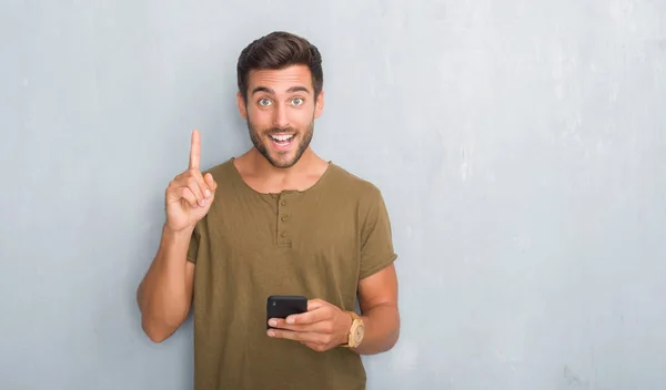 英俊的年轻人在灰色垃圾墙上发短信使用智能手机惊讶的想法或问题指向手指与快乐的脸 — 图库照片