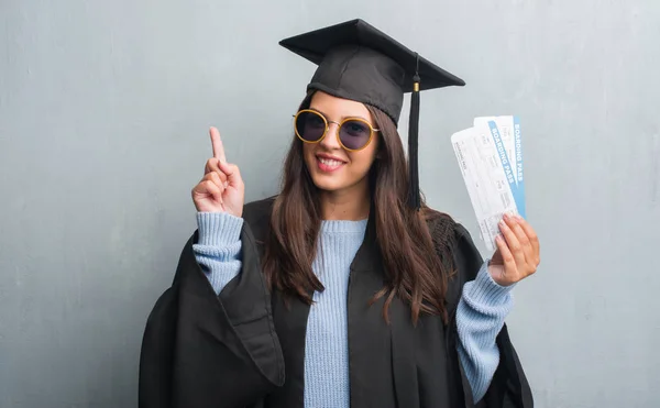 グランジ灰色の壁を身に着けている卒業の若いブルネットの女性アイデアに驚いて搭乗券を保持している制服や幸せそうな顔 ナンバーワンと人差し指の質問 — ストック写真