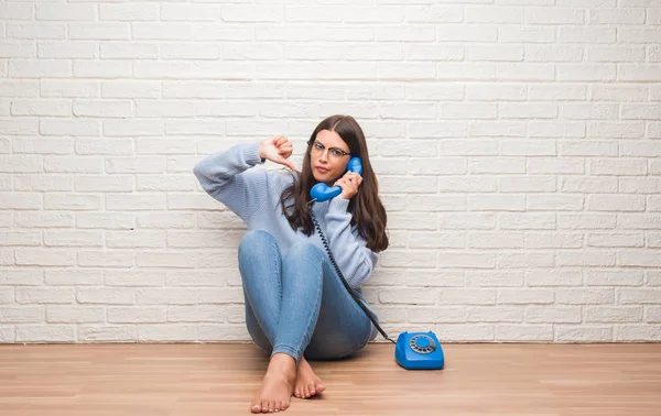 Young brunette woman sitting on the floor calling on vintage telephone with angry face, negative sign showing dislike with thumbs down, rejection concept