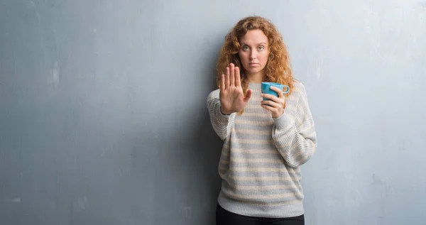 灰色垃圾墙上的年轻红头发的女人喝一杯咖啡用张开手做停止标志以严肃和自信的表达 防御姿态 — 图库照片