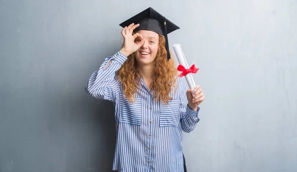 指を通して見る目に手で サインをして笑っている幸せそうな顔で灰色のグランジ壁持株ディプロマを卒業した若い赤毛の女性 — ストック写真