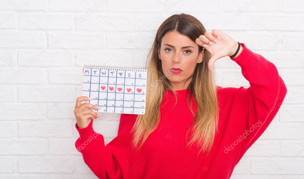 Young adult woman over white brick wall holding period calendar with angry face, negative sign showing dislike with thumbs down, rejection concept