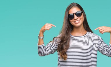 Genç güzel Arap kadın yüzünde gülümseme ile kendinden emin görünüyorsun, kendini gururlu ve mutlu parmakları ile işaret izole arka plan üzerinde güneş gözlüğü takıyor.