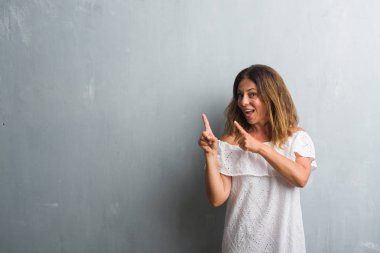 Orta yaş İspanyol kadın gülümseyerek ve iki elleri ve parmakları ile tarafına işaret kamera bakarak gri grunge duvarın üzerinden ayakta.