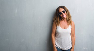 Orta yaş İspanyol kadın dilini güneş gözlüğü takıyor gri duvarın üzerinden out komik ifade ile mutlu. Duygu kavramı.