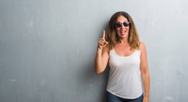 Orta yaş İspanyol kadın gri Duvar ile başarılı fikir parmak işaret güneş gözlüğü takıyor. Çıktıktan ve mutlu. Bir numara.