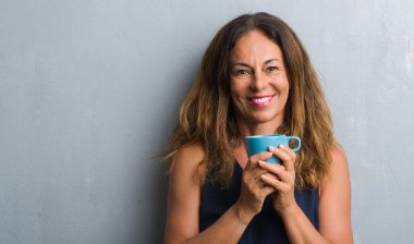 Duran duran ve diş gösterilen kendine güvenen bir gülümsemeyle gülümseyerek mutlu bir yüz ile kahve içme gri grunge duvarın üzerinden orta yaş İspanyol kadın