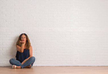 Orta yaş İspanyol kadın bir öpücük el ile güzel ve seksi olmak hava üfleme kamera bakıyor beyaz tuğla duvarın üzerinden yere oturma. Aşk ifade.