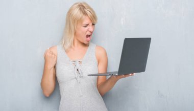 Yetişkin beyaz kadın bilgisayar dizüstü kullanarak grunge gri duvar yüzünden rahatsız ve boşa çıkarmak ile öfke, çılgın da yükseltilmiş el ile bağırmaya kavramı öfke