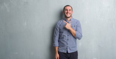 Genç beyaz adam gri grunge duvarın neşeli bir gülümseme ile el ve parmak kadar kameraya bakarak yüzündeki mutlu ve doğal ifade ile yan ile işaret yüz üzerinden.