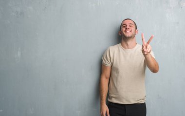 Beyaz delikanlı zafer işareti yapıyor parmak gösterilen kamera seyir gülümseyen gri grunge duvarın üzerinden. İki numaralı.