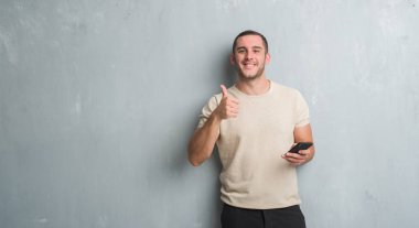 Gri grunge Genç beyaz adam manifatura smartphone mutlu ok işareti yapıyor büyük gülümseme ile kullanarak bir ileti duvar, parmaklarıyla, mükemmel işaret parmak